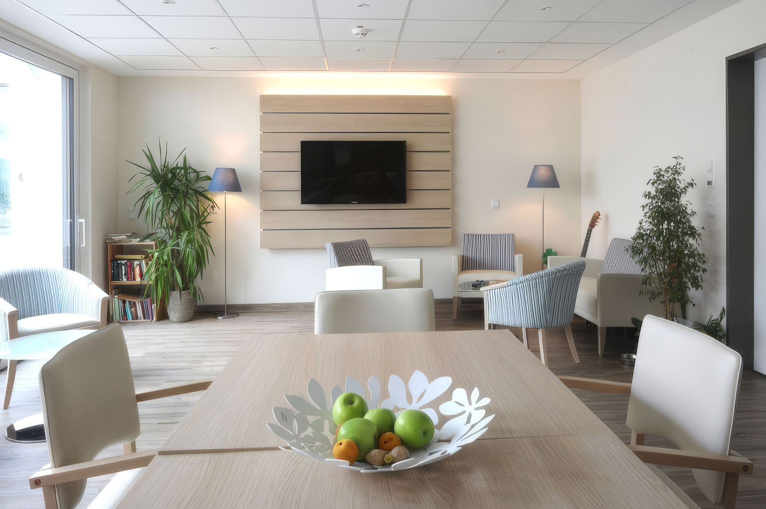 Heller Gemeinschaftsraum im Hospiz: Holztisch mit Obstschale, helle Sessel, Pflanzen und ein Bücherregal vor großen Fenstern